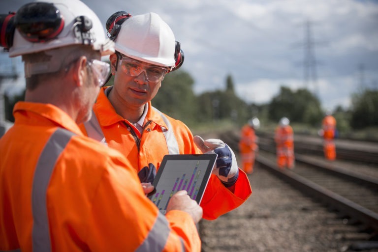 Two construction workers in hard hats looking at tablet standing next to train tracks 
