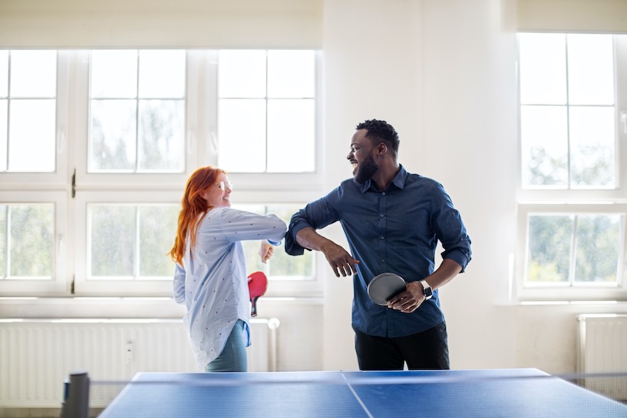 Corporate wellness program colleagues giving elbow bump while playing table tennis in office