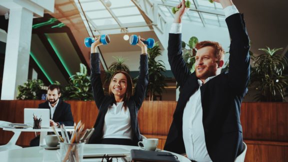 Two employees lifting weights over their heads in corporate center | Holistic Employee Wellness | Limeade