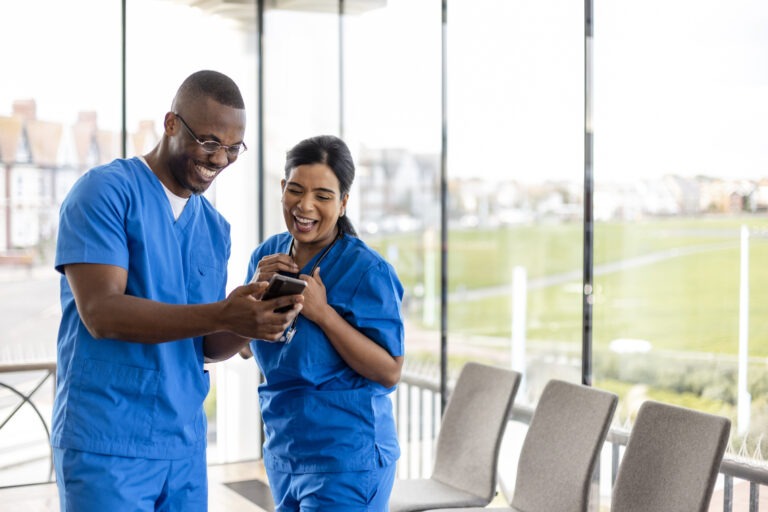 Mixed ethnic group of medical professionals laughing together while receiving an employee communication on their phone. They are working a shift at a hospital and are dressed in scrubs.