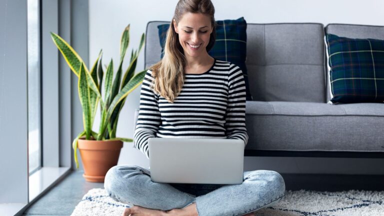 Smiling woman sits on her living room floor using silver laptop in lap | Employee Mental Health