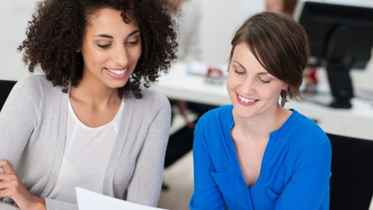 Two female, engaged employees smile while looking at paper report | Organizational Culture